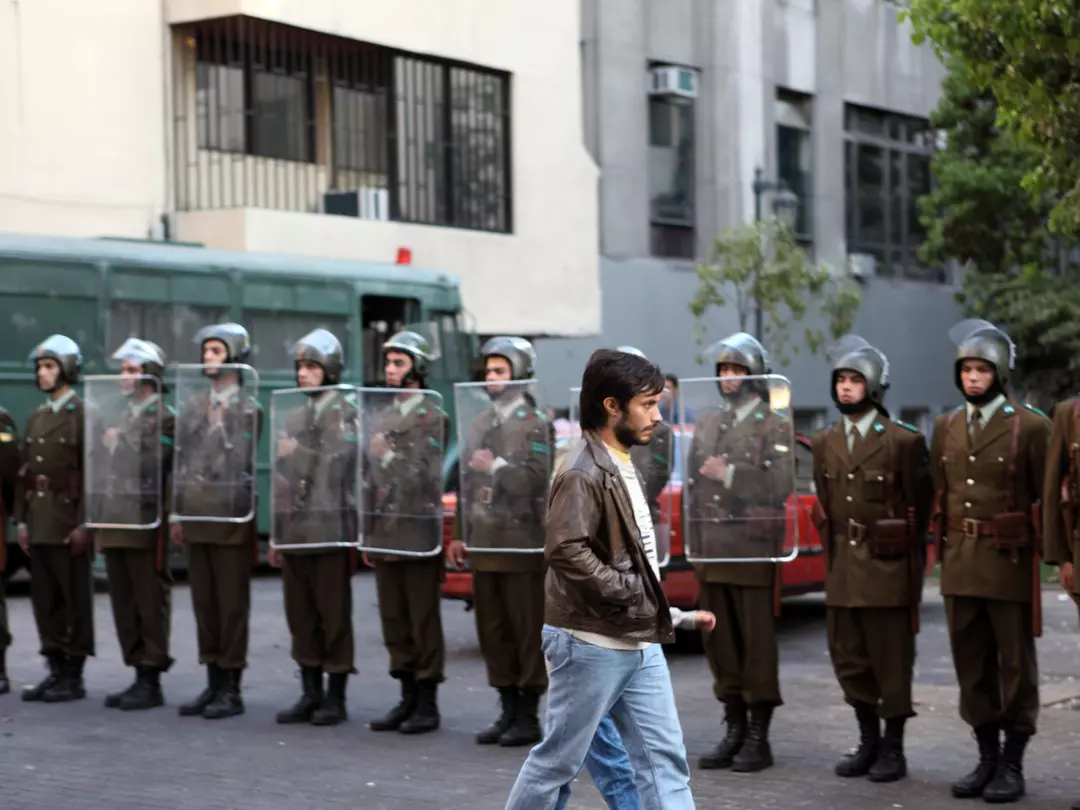 Bernal walking in front of a line of army personnel dressed in riot gear.