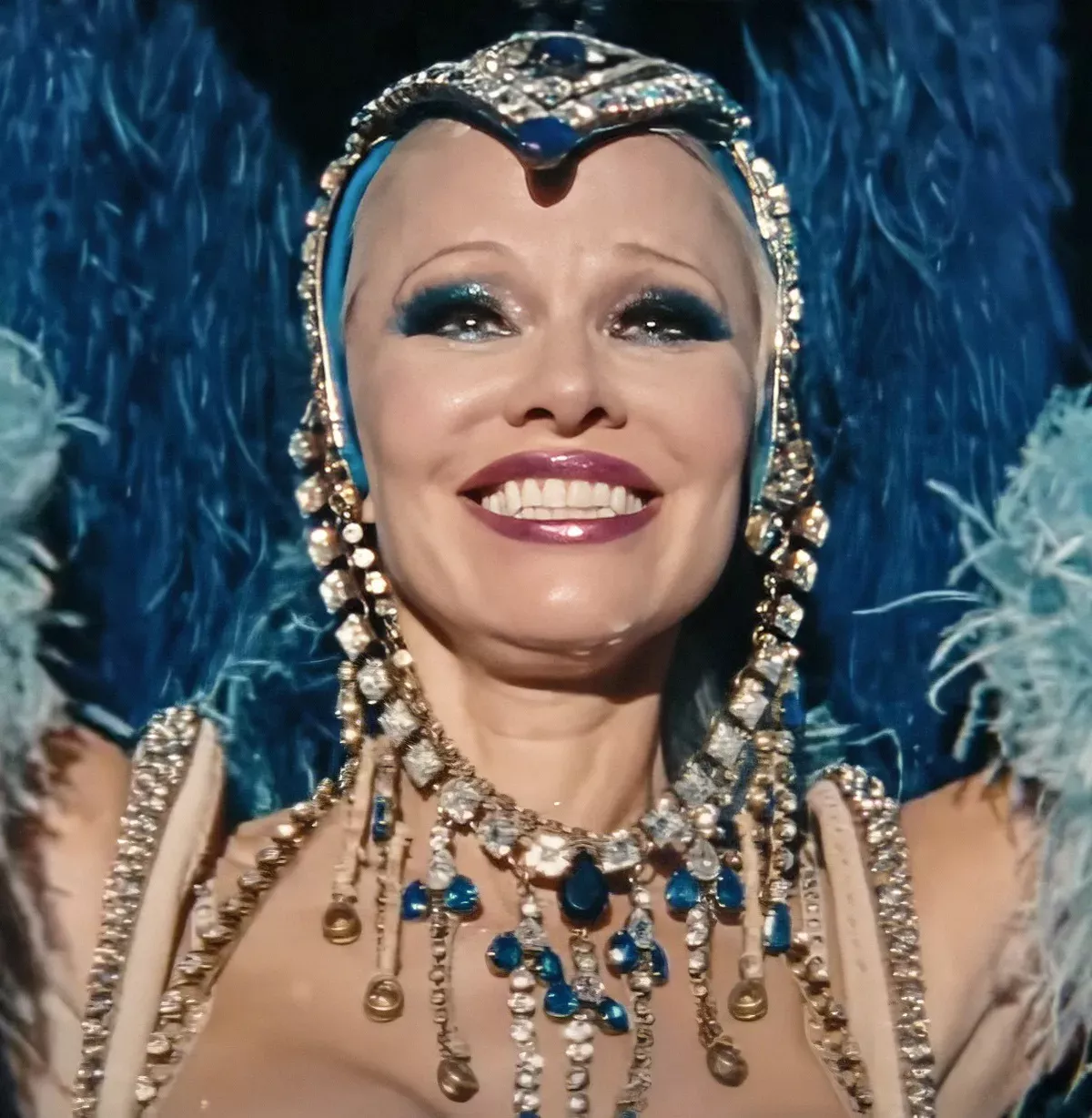 A closeup of a showgirl in a blue head dress. She is smiling.