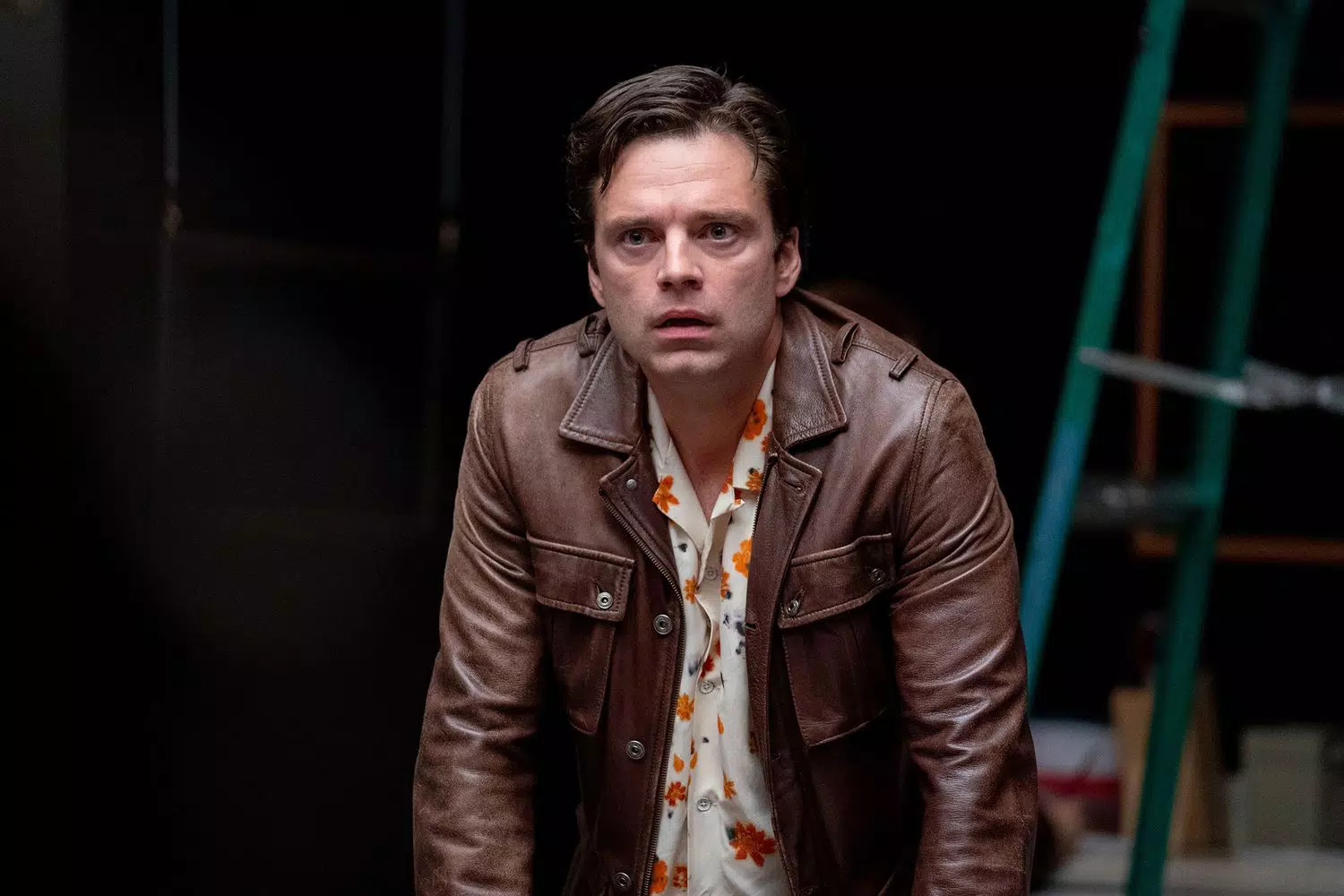 Sebastian Stan, wearing a brown leather jacket and a floral shirt, looking stunned, in A Different Man