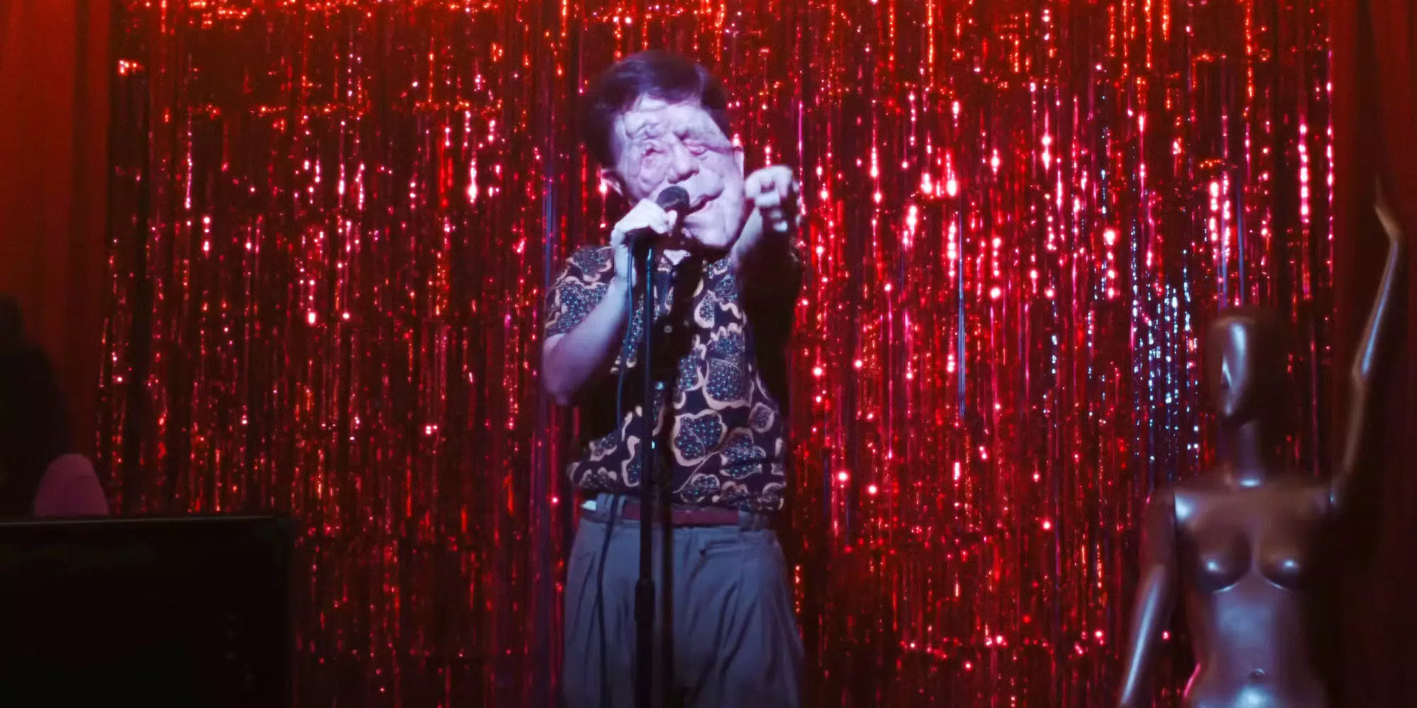 Oswald in A Different Man. He is signing into a microphone with a red, glittery background.