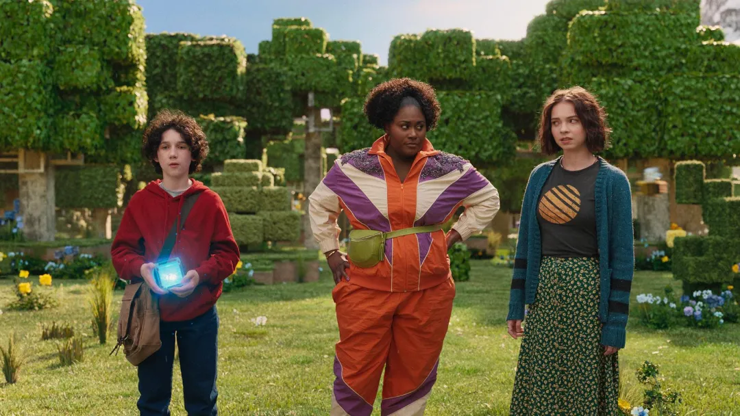 A young boy, a woman, and a teenage girl stand in a green clearing. The boy is holding a glowing blue cube
