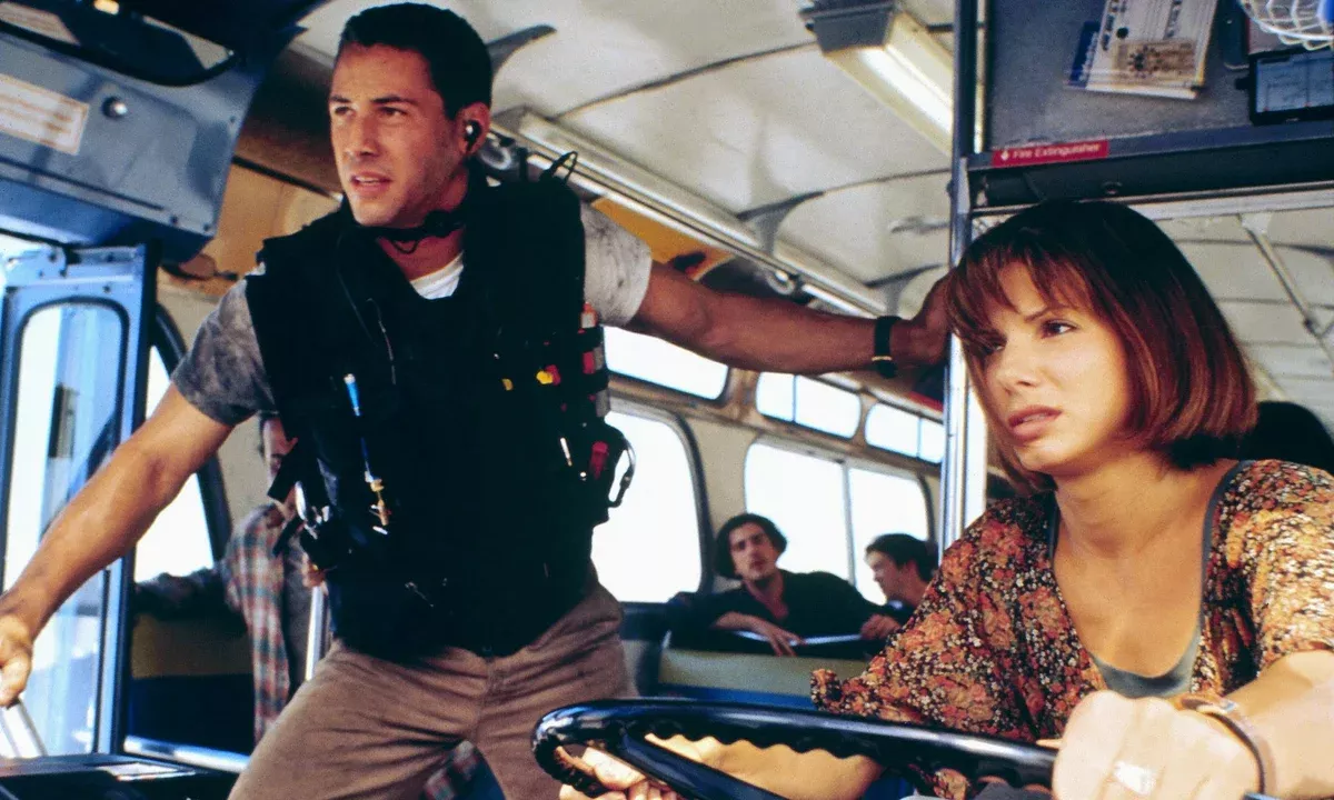 a man in a bulletproof stands on a bus, next to a female bus driver