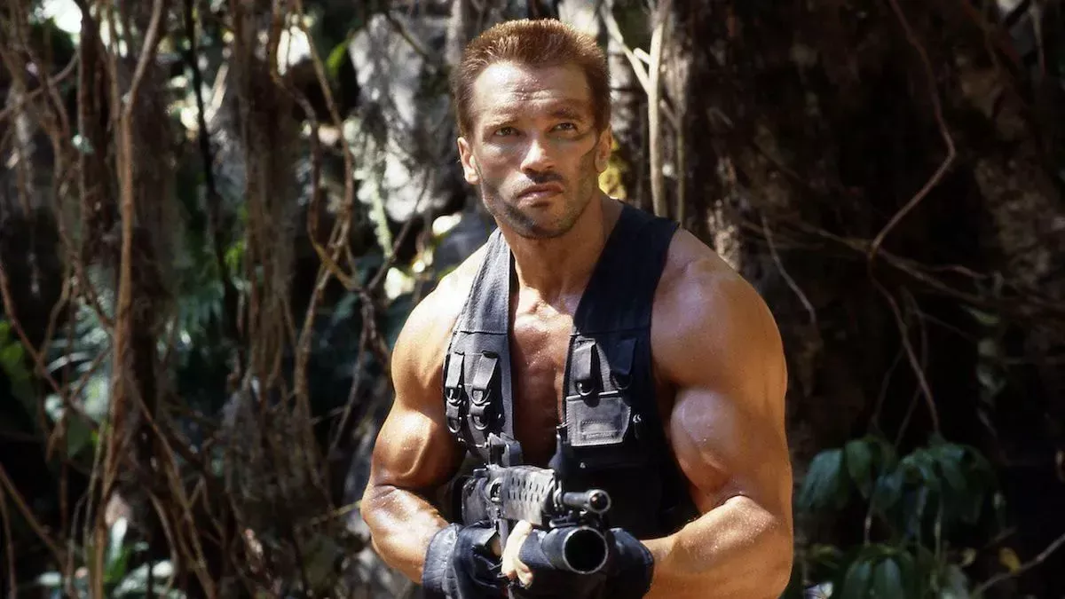 a large muscled man holds a machine gun in the jungle