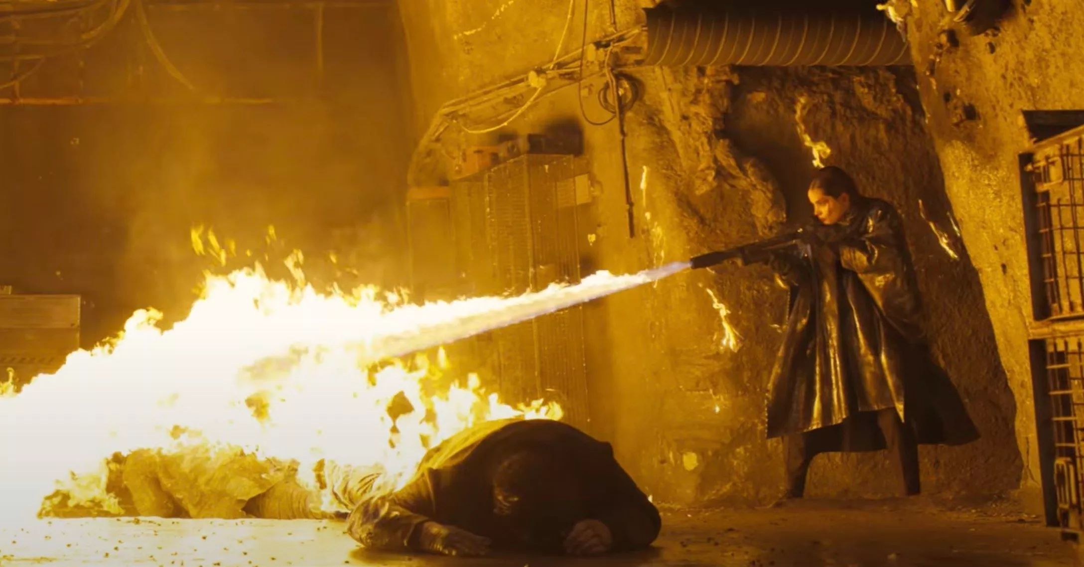 a woman in a black trenchcoat uses a giant flamethrower